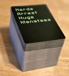 A large stack of playing cards. The top card is black with green writing that says "Nerds Arrest Huge Manatees"