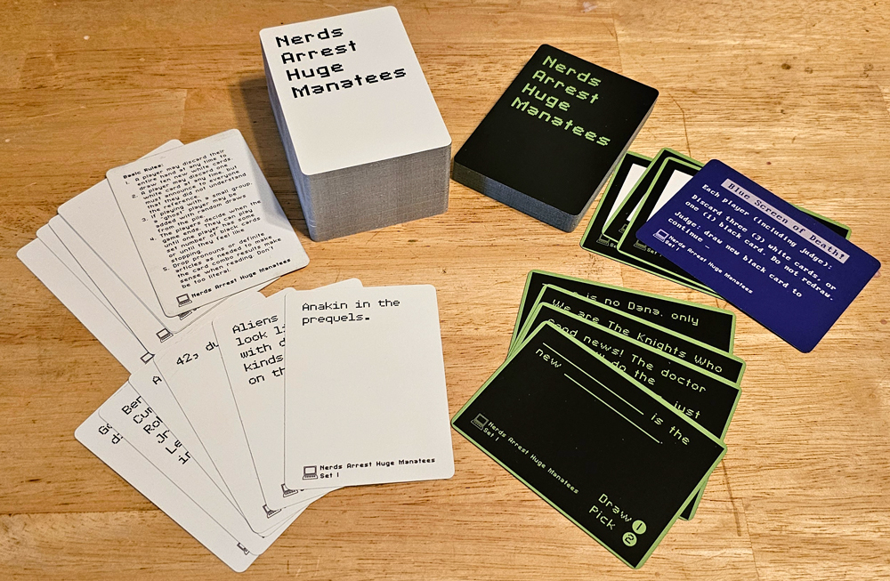 Stacks and spreads of white and black cards. The backs read "Nerds Arrest Huge Manatees." The fronts say things like "Anakin in the prequels." and "__________ is the new ________." One blue card is the Blue Screen of Death and includes the rules for that card in the game.