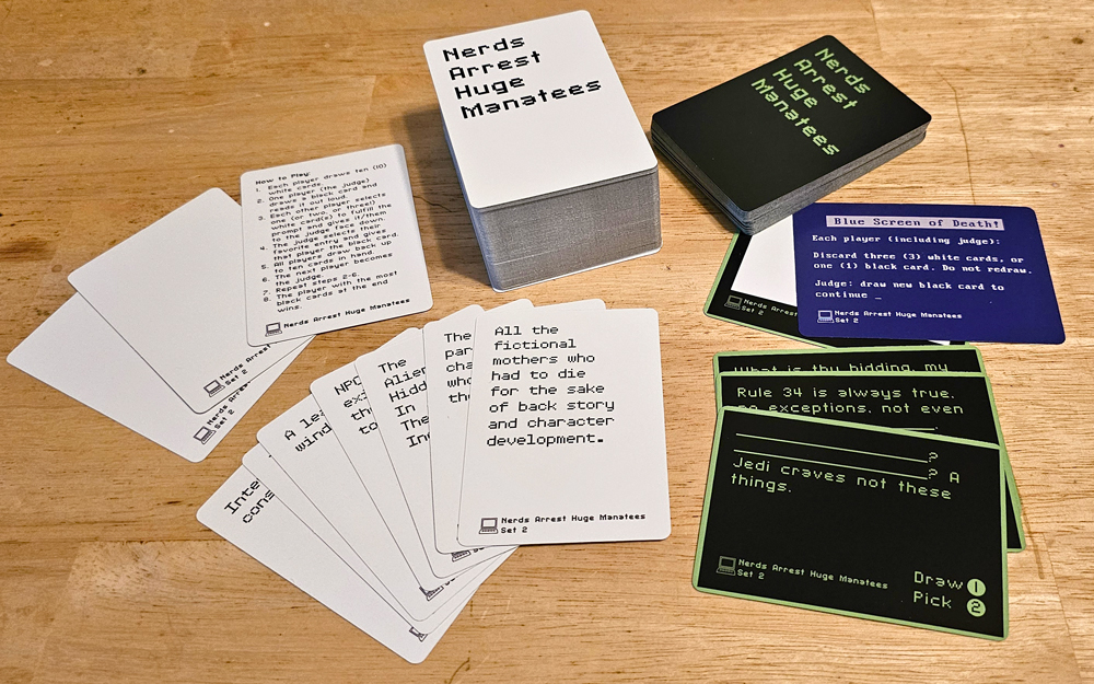 Stacks and spreads of white and black cards. The backs read "Nerds Arrest Huge Manatees." The fronts say things like "All the fictional mothers who had to die for the sake of back story and character development." and "__________? _________? a Jedi craves not these things." One blue card is the Blue Screen of Death and includes the rules for that card in the game.