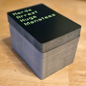 A large stack of playing cards. The top card is black with green writing that says "Nerds Arrest Huge Manatees"