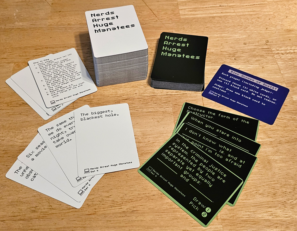 Stacks and spreads of white and black cards. The backs read "Nerds Arrest Huge Manatees." The fronts say things like "The biggest, blackest hole." and "In the criminal justice system, the people are represented by two separate yet equally important groups: _____ and _____."